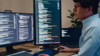 Homem de traços asiáticos, usando óculos e camisa, trabalha com duas telas de computador. Nelas, podemos ver códigos de uma linguagem de programação não identificada.