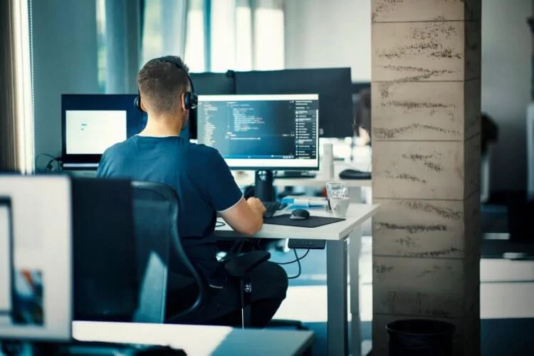 Homem de costas olhando para a tela de um computador enquanto digita códigos de linguagem de programação. Ele veste uma camiseta azul e usa fones de ouvido. O ambiente, que parece um escritório, tem outros computadores.