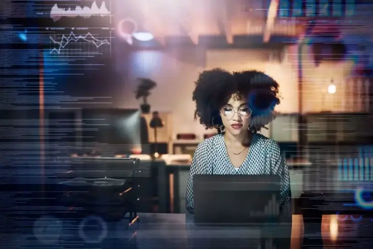 Mulher negra de óculos digitando códigos de programação em uma tela de computador. Ela parece estar em um ambiente de escritório. Sobreposta à imagem dela há linhas e gráficos.