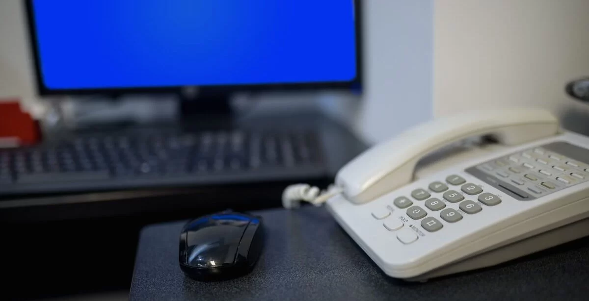 Um telefone branco sobre uma mesa de escritório. Há um mouse preto perto dele e um teclado com uma tela azul ao fundo. Possivelmente, são equipamentos de um escritório.