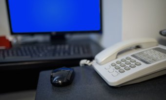 Um telefone branco sobre uma mesa de escritório. Há um mouse preto perto dele e um teclado com uma tela azul ao fundo. Possivelmente, são equipamentos de um escritório.