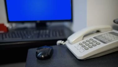 Um telefone branco sobre uma mesa de escritório. Há um mouse preto perto dele e um teclado com uma tela azul ao fundo. Possivelmente, são equipamentos de um escritório.