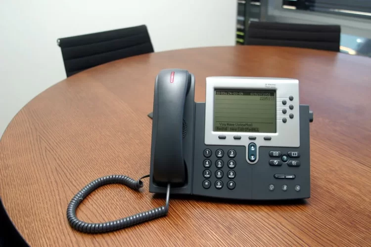 Telefone cinza e preto com sistema de PABX. Ele aparece sobre uma mesa de madeira, possivelmente a sala de reunião de um escritório. Há algumas cadeiras vazias ao redor da mesa. 