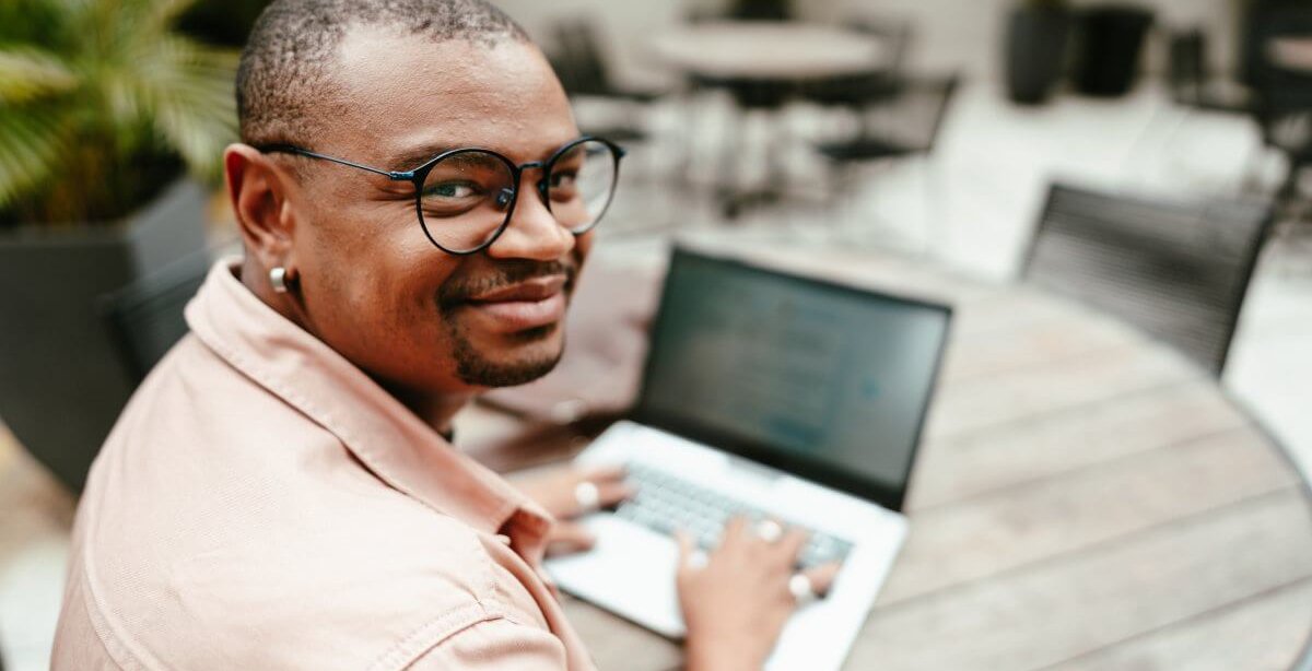 Um homem negro de óculos usando camisa social está digitando informações em um notebook. Ele aparentemente está em um café durante o dia, e há outras mesas ao fundo para complementar o ambiente.
