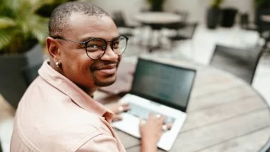 Um homem negro de óculos usando camisa social está digitando informações em um notebook. Ele aparentemente está em um café durante o dia, e há outras mesas ao fundo para complementar o ambiente.