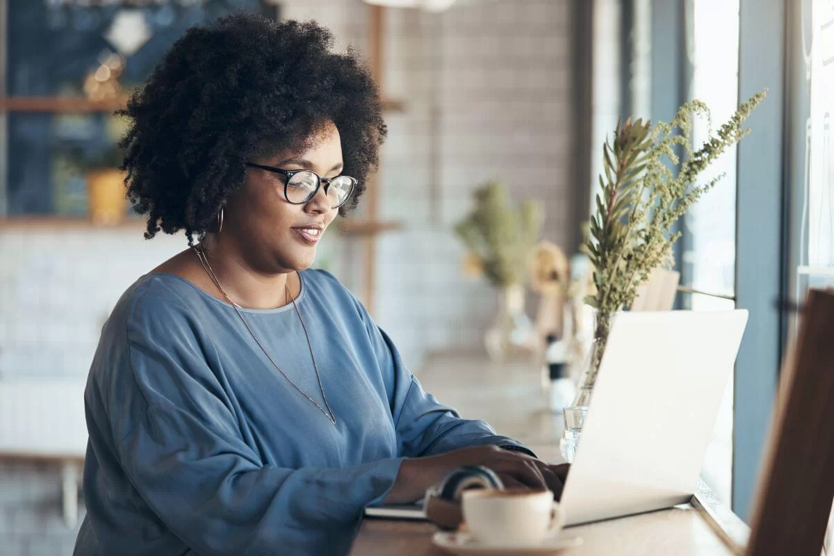 Mulher negra com óculos e blusa azul sentada digitando em um teclado de notebook. Perto dela há uma xícara de café e um vaso com plantas. Ela parece estar trabalhando em uma cafeteria. 