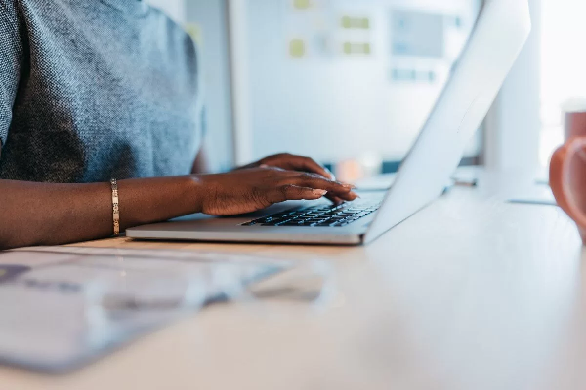 Mulher negra digitando URL em um teclado de notebook. Ela usa uma roupa em tom de azul. O rosto dela não aparece. 