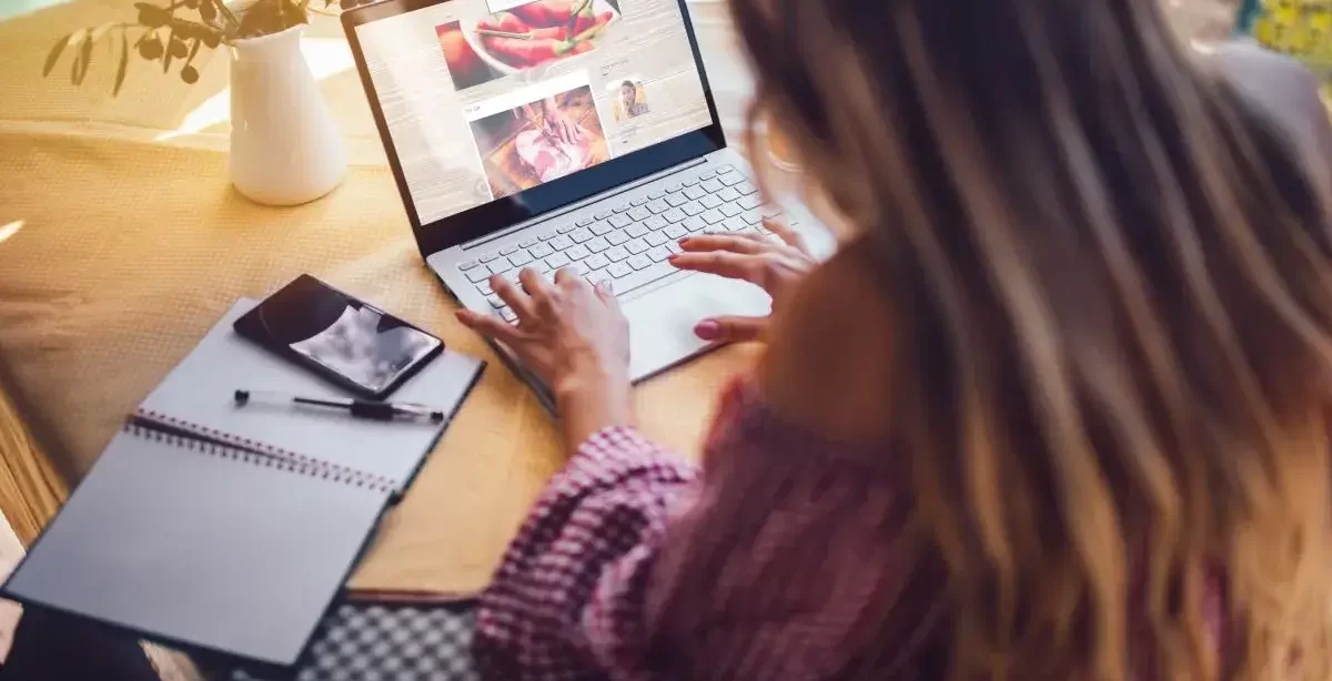 A imagem mostra uma mulher de costas para a câmera, trabalhando em um notebook. Ela está editando um blog de culinária e, para isso, precisa de um CMS. Ao redor dela, sobre a mesa, há um caderno aberto, uma caneta e um celular. Atrás do computador temos um vaso de flores.