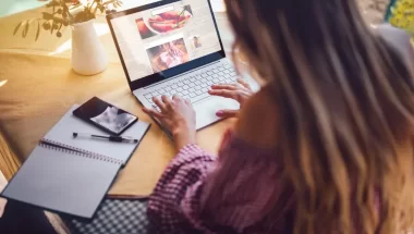 A imagem mostra uma mulher de costas para a câmera, trabalhando em um notebook. Ela está editando um blog de culinária e, para isso, precisa de um CMS. Ao redor dela, sobre a mesa, há um caderno aberto, uma caneta e um celular. Atrás do computador temos um vaso de flores.