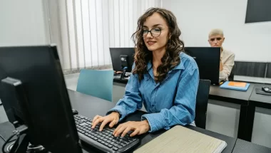A imagem mostra uma mulher de óculos sentada de frente para um computador. Ela está com uma camisa azul e digita algo no teclado. Há outra pessoa na cadeira ao fundo, completando o cenário.
