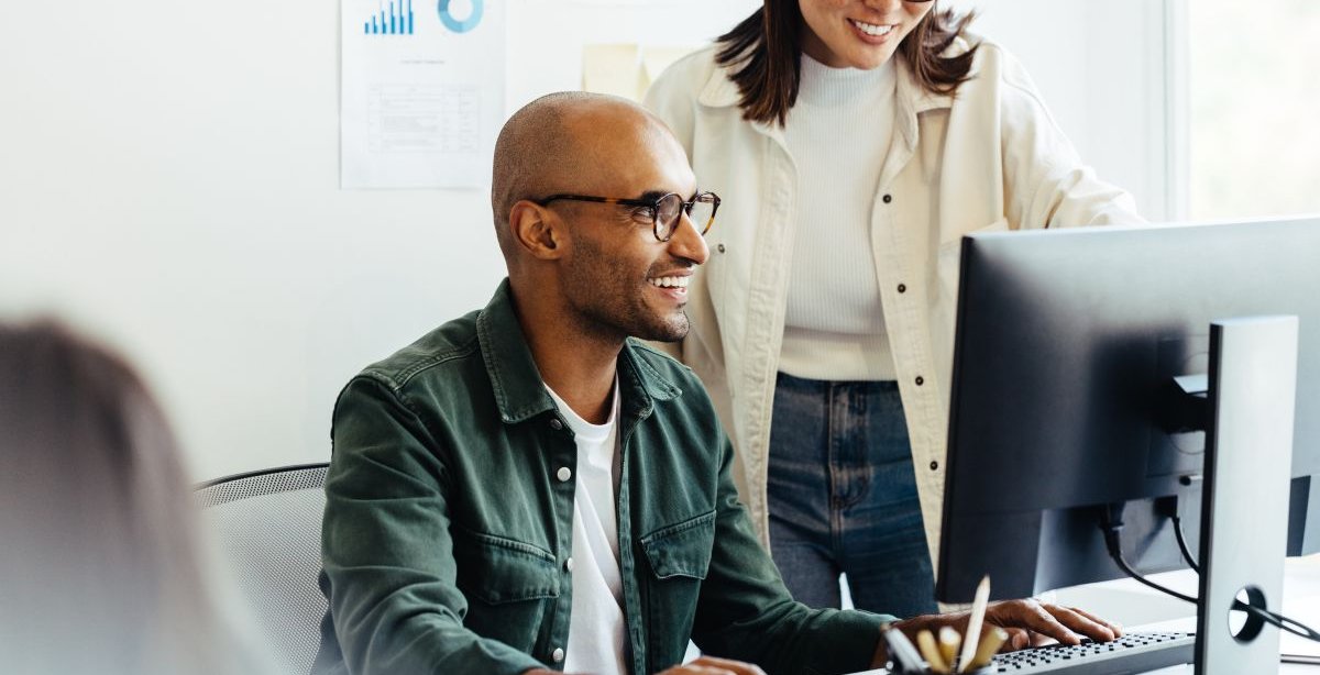 A imagem mostra um homem e uma mulher conversando em um escritório. Eles vestem blusas e usam óculos, e estão falando sobre o desenvolvimento de um layout de site. Outra pessoa aparece desfocada na imagem, mas ela não participa da conversa.