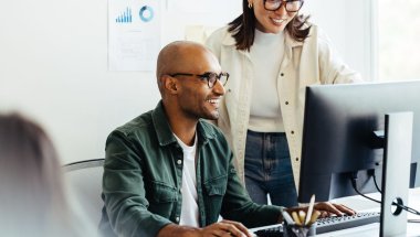 A imagem mostra um homem e uma mulher conversando em um escritório. Eles vestem blusas e usam óculos, e estão falando sobre o desenvolvimento de um layout de site. Outra pessoa aparece desfocada na imagem, mas ela não participa da conversa.