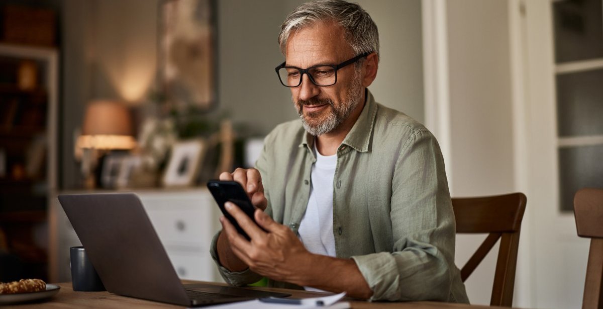 A imagem mostra um homem de meia-idade diante de um notebook e com um celular na mão. Após alguns trabalhos com localhost, ele verifica o funcionamento de uma aplicação no smartphone.
