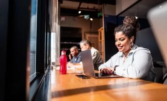 A imagem mostra uma mulher em um escritório diante de uma janela, trabalhando em otimização de sites. Ela está tocando o touchpad do notebook. Ao fundo, é possível ver mais três pessoas trabalhando no mesmo ambiente.