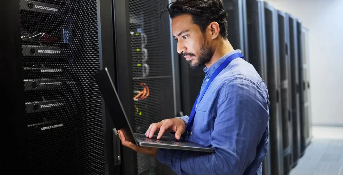 A imagem mostra um homem de barba vestindo uma camisa social azul e segurando um notebook. Ele está analisando a infraestrutura de servidor web de uma empresa, em uma sala com diversos servidores instalados.