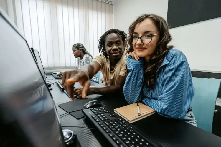 A imagem mostra um homem negro conversando sobre como programar em SQL com uma moça branca de óculos e cabelos longos. Eles estão diante de um computador em uma mesa de escritório, e outra colega de trabalho aparece sentada ao fundo na imagem.