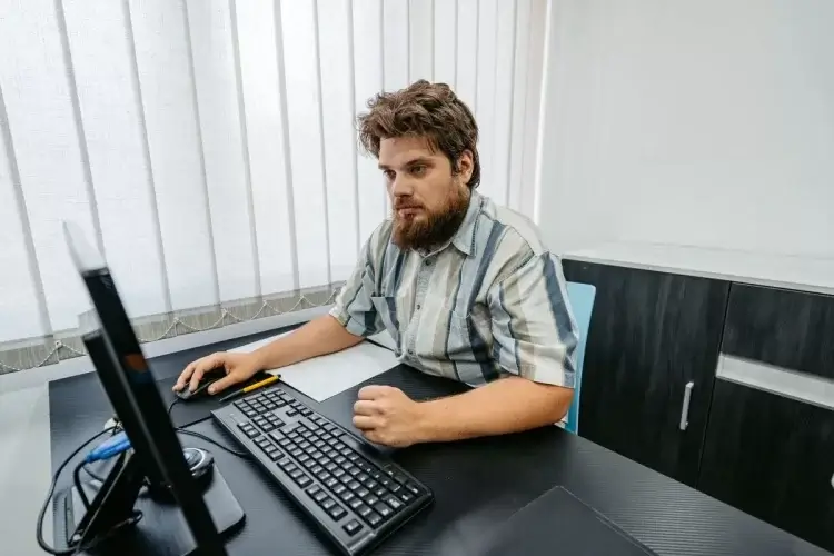 A imagem mostra um homem branco, jovem e de barba em um escritório. Ele está diante de um computador, programando com auxílio do SQL.