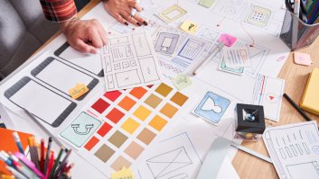 Aparentemente, duas pessoas da área de marketing estão trabalhando em um projeto de uma landing page. Eles estão criando um esboço inicial para definir a estrutura e o layout. Não aparecem os rostos deles, apenas as mãos.