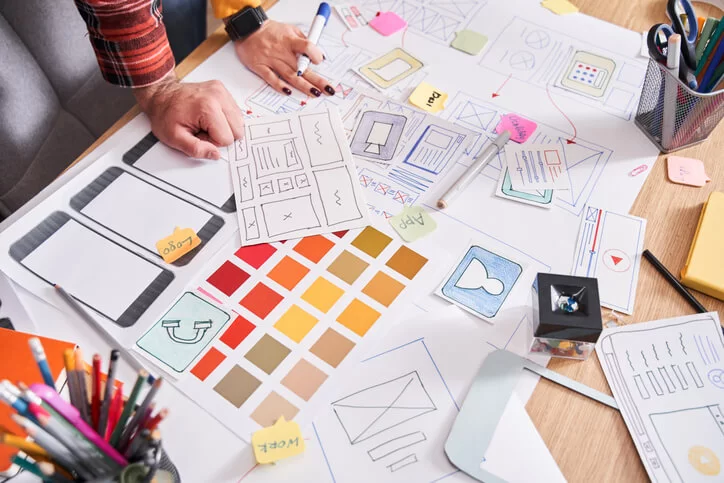 Aparentemente, duas pessoas da área de marketing estão trabalhando em um projeto de uma landing page. Eles estão criando um esboço inicial para definir a estrutura e o layout. Não aparecem os rostos deles, apenas as mãos.