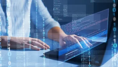 A imagem mostra um homem digitando códigos de programação em um notebook. O rosto dele não aparece. Podemos ver o recurso de IA para programação em ação, já que aparecem vários códigos binários e outros códigos em primeiro plano.