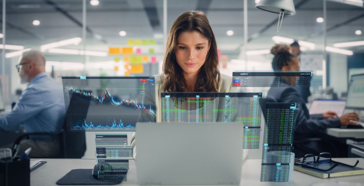 A imagem mostra uma mulher em um escritório, rodeada por colegas de trabalho. Na mesa dela há um notebook, onde ela usa a Inteligência Artificial para escrever textos, enquanto analisa gráficos e outros dados referentes ao conteúdo.