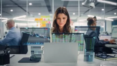 A imagem mostra uma mulher em um escritório, rodeada por colegas de trabalho. Na mesa dela há um notebook, onde ela usa a Inteligência Artificial para escrever textos, enquanto analisa gráficos e outros dados referentes ao conteúdo.