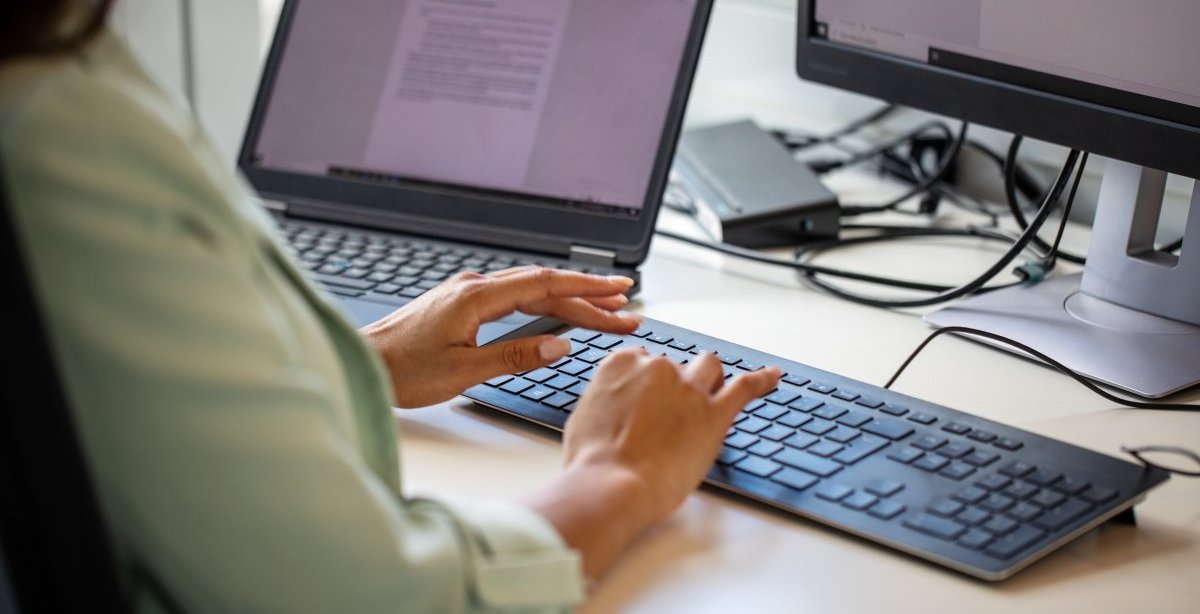 A imagem mostra uma mulher digitando em um teclado de computador em uma mesa de escritório. O rosto dela não aparece. Ela tem um notebook com um documento aberto ao lado, no qual checa um texto que foi reescrito com a Inteligência Artificial do ChatGPT.