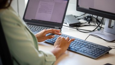 A imagem mostra uma mulher digitando em um teclado de computador em uma mesa de escritório. O rosto dela não aparece. Ela tem um notebook com um documento aberto ao lado, no qual checa um texto que foi reescrito com a Inteligência Artificial do ChatGPT.