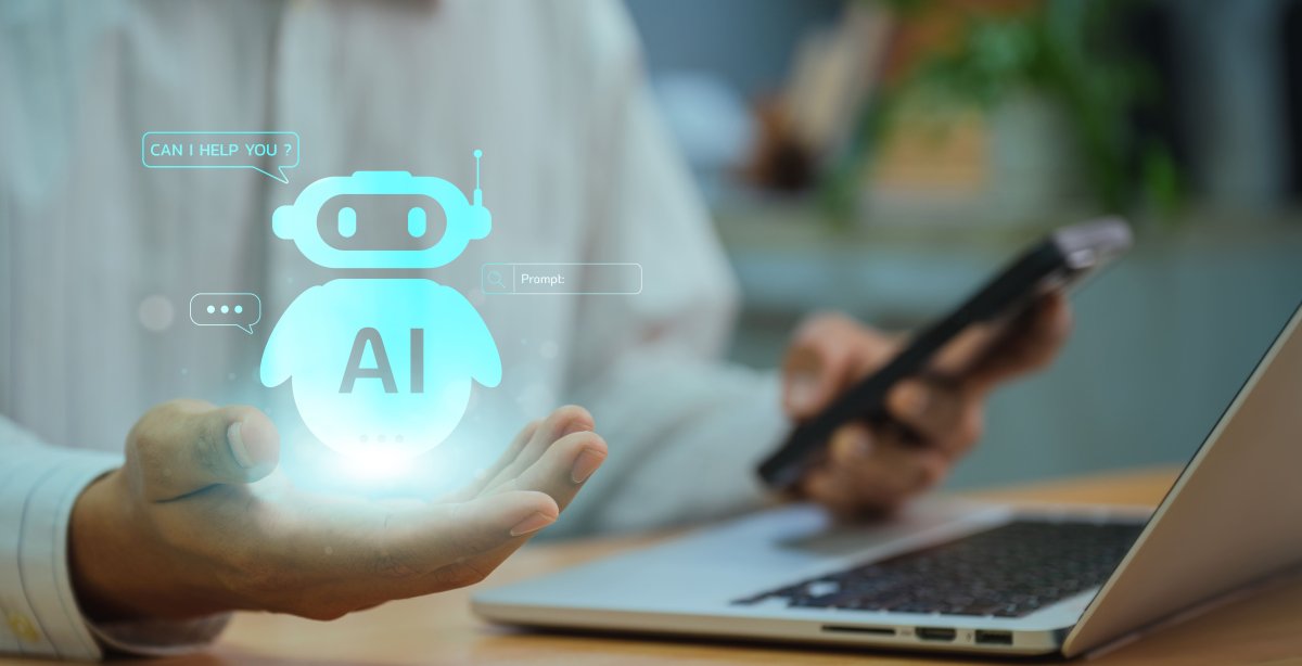 Uma pessoa segura um smartphone com a mão direita enquanto a mão esquerda paira sobre o teclado de um laptop. Um ícone holográfico de robô AI com uma mensagem dizendo "POSSO AJUDÁ-LO?" está flutuando acima da palma da mão esquerda.
