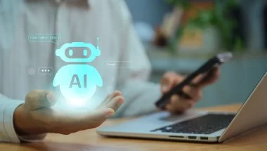 Uma pessoa segura um smartphone com a mão direita enquanto a mão esquerda paira sobre o teclado de um laptop. Um ícone holográfico de robô AI com uma mensagem dizendo "POSSO AJUDÁ-LO?" está flutuando acima da palma da mão esquerda.