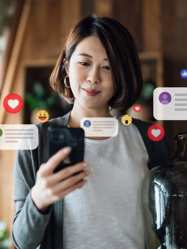 Mulher oriental segura um smartphone com a mão direita. A pessoa em questão usa roupas casuais e está em um ambiente que parece um café, vendo suas redes sociais. Da tela do aparelho surgem ícones que simulam interações de redes sociais.