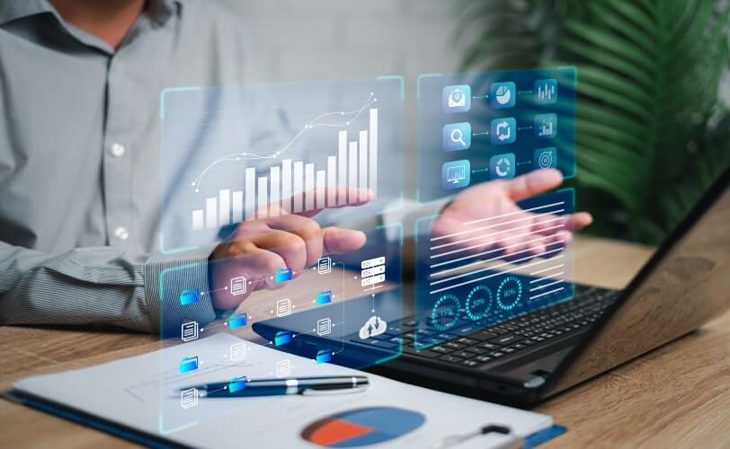A imagem mostra uma pessoa digitando em um teclado, enquanto gráficos e painéis analíticos flutuam em forma de hologramas sobre a mesa. Os elementos visuais incluem barras de crescimento, gráficos circulares e indicadores de desempenho, simbolizando o uso de ferramentas digitais e de inteligência de dados no ambiente corporativo. A cena representa o poder da transformação digital, em que dados e tecnologia se unem para gerar insights estratégicos, apoiar a tomada de decisões baseadas em evidências e otimizar processos empresariais.