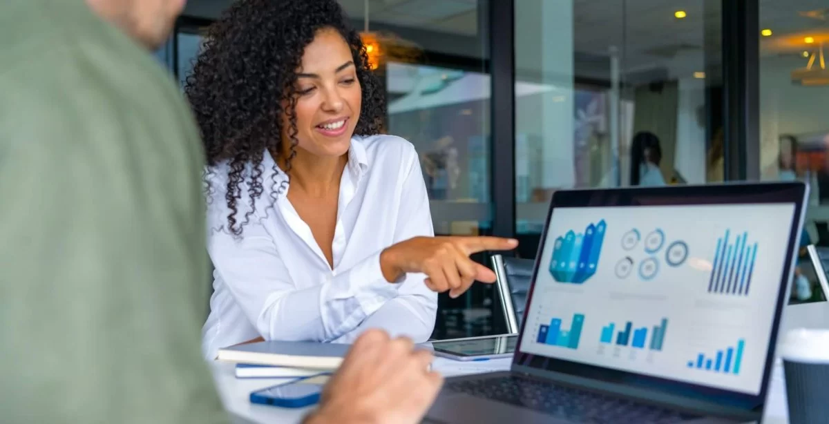 A imagem mostra duas pessoas em uma reunião de negócios, focadas em uma tela de laptop com gráficos e indicadores de desempenho. A mulher à direita sorri e aponta para os dados, indicando um momento de discussão e tomada de decisão baseada em informações analíticas. O ambiente corporativo e moderno reforça a importância da inteligência de dados e da comunicação eficaz no ambiente de trabalho, evidenciando como a análise visual e a tecnologia impulsionam estratégias e resultados nas empresas.