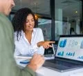 A imagem mostra duas pessoas em uma reunião de negócios, focadas em uma tela de laptop com gráficos e indicadores de desempenho. A mulher à direita sorri e aponta para os dados, indicando um momento de discussão e tomada de decisão baseada em informações analíticas. O ambiente corporativo e moderno reforça a importância da inteligência de dados e da comunicação eficaz no ambiente de trabalho, evidenciando como a análise visual e a tecnologia impulsionam estratégias e resultados nas empresas.