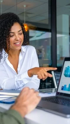 A imagem mostra duas pessoas em uma reunião de negócios, focadas em uma tela de laptop com gráficos e indicadores de desempenho. A mulher à direita sorri e aponta para os dados, indicando um momento de discussão e tomada de decisão baseada em informações analíticas. O ambiente corporativo e moderno reforça a importância da inteligência de dados e da comunicação eficaz no ambiente de trabalho, evidenciando como a análise visual e a tecnologia impulsionam estratégias e resultados nas empresas.