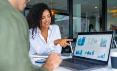 A imagem mostra duas pessoas em uma reunião de negócios, focadas em uma tela de laptop com gráficos e indicadores de desempenho. A mulher à direita sorri e aponta para os dados, indicando um momento de discussão e tomada de decisão baseada em informações analíticas. O ambiente corporativo e moderno reforça a importância da inteligência de dados e da comunicação eficaz no ambiente de trabalho, evidenciando como a análise visual e a tecnologia impulsionam estratégias e resultados nas empresas.