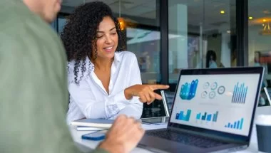 A imagem mostra duas pessoas em uma reunião de negócios, focadas em uma tela de laptop com gráficos e indicadores de desempenho. A mulher à direita sorri e aponta para os dados, indicando um momento de discussão e tomada de decisão baseada em informações analíticas. O ambiente corporativo e moderno reforça a importância da inteligência de dados e da comunicação eficaz no ambiente de trabalho, evidenciando como a análise visual e a tecnologia impulsionam estratégias e resultados nas empresas.