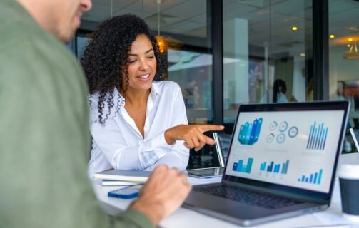 A imagem mostra duas pessoas em uma reunião de negócios, focadas em uma tela de laptop com gráficos e indicadores de desempenho. A mulher à direita sorri e aponta para os dados, indicando um momento de discussão e tomada de decisão baseada em informações analíticas. O ambiente corporativo e moderno reforça a importância da inteligência de dados e da comunicação eficaz no ambiente de trabalho, evidenciando como a análise visual e a tecnologia impulsionam estratégias e resultados nas empresas.