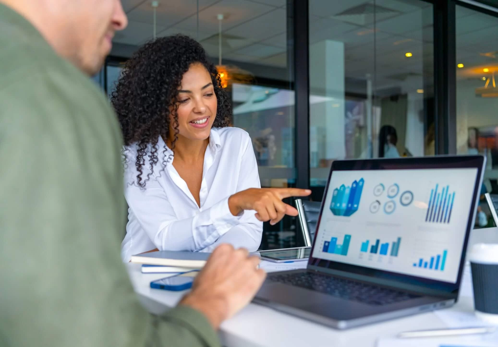 A imagem mostra duas pessoas em uma reunião de negócios, focadas em uma tela de laptop com gráficos e indicadores de desempenho. A mulher à direita sorri e aponta para os dados, indicando um momento de discussão e tomada de decisão baseada em informações analíticas. O ambiente corporativo e moderno reforça a importância da inteligência de dados e da comunicação eficaz no ambiente de trabalho, evidenciando como a análise visual e a tecnologia impulsionam estratégias e resultados nas empresas.
