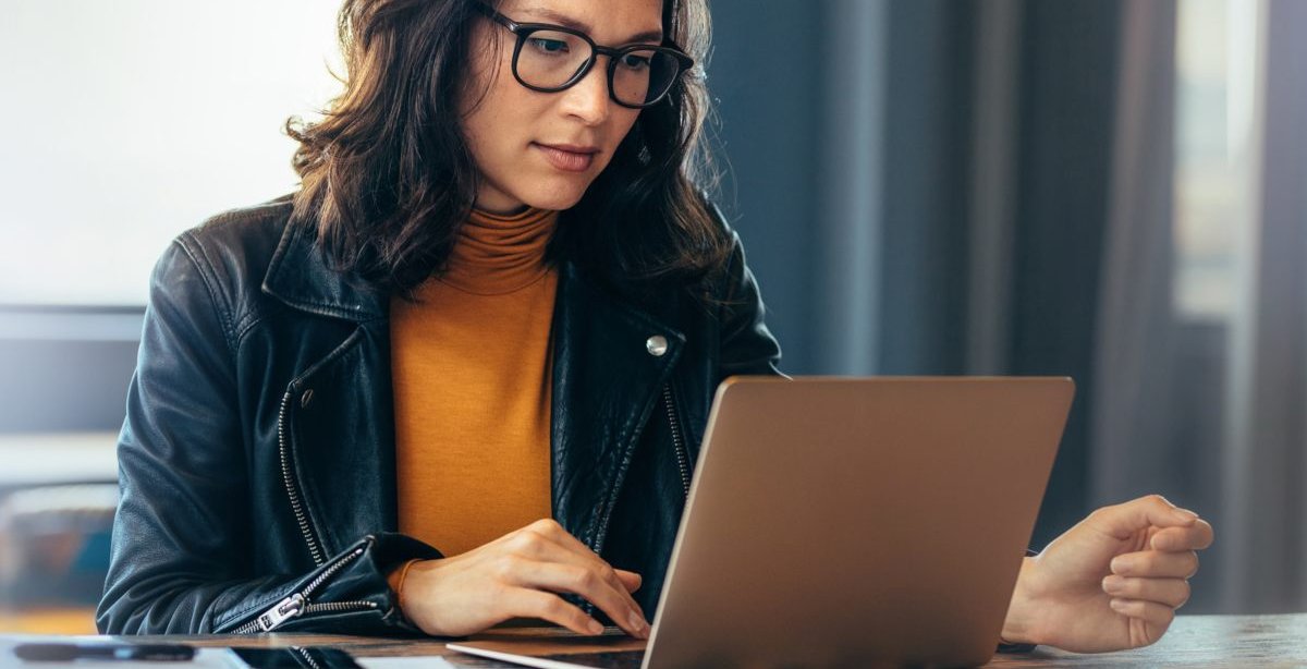 A imagem mostra uma mulher digitando em um notebook, provavelmente em um escritório. Ela está fazendo uma pesquisa para obter informações sobre FQDN, já que ele é importante para o funcionamento de uma rede.