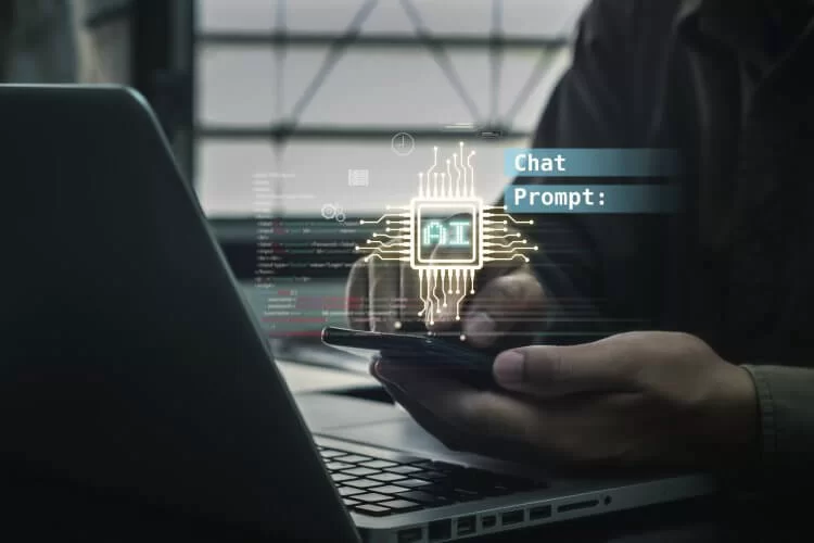 A foto mostra um homem de negócios em frente a um notebook, interagindo com um Chat de IA em um celular. Não aparece o rosto dele. Ao fundo podemos ver algumas linhas de interações e à frente, em destaque, as palavras Chat e Prompt