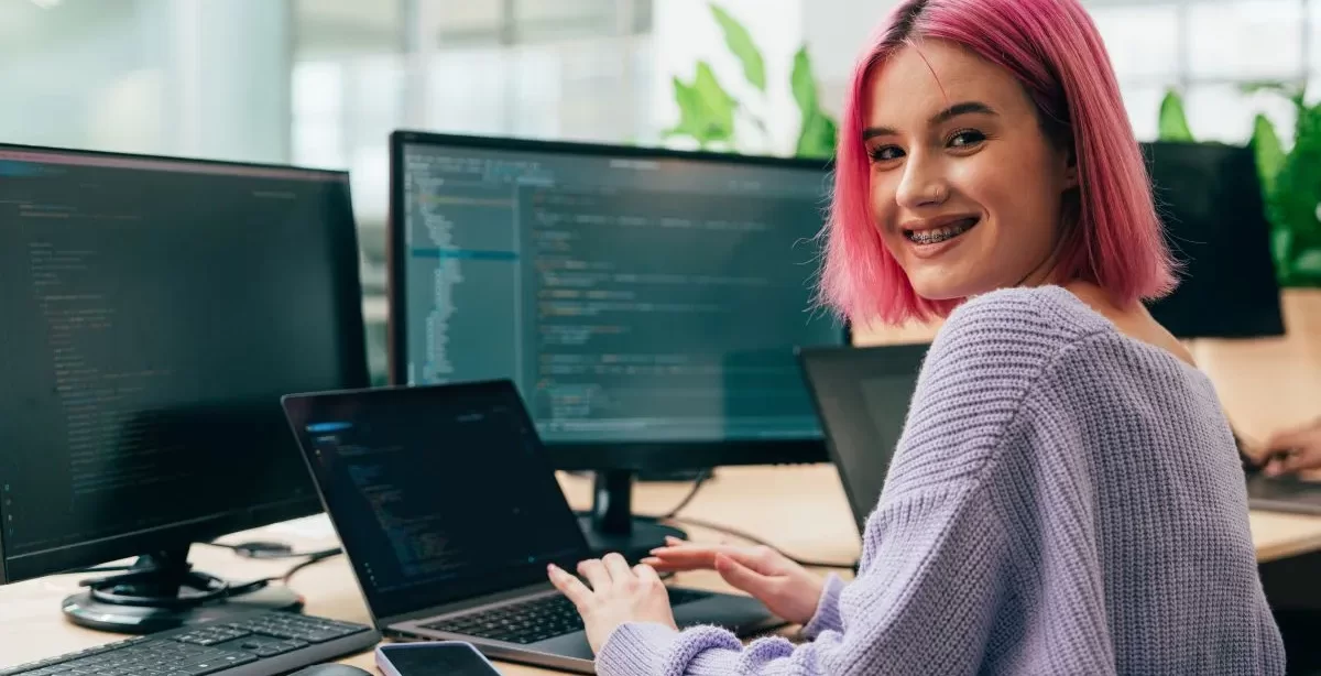 A imagem mostra uma jovem programadora branca de cabelos cor-de-rosa. Ela está trabalhando em uma empresa e os monitores que estão à sua frente mostram várias linhas de código de programação feitas com auxílio de Python.
