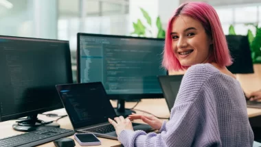 A imagem mostra uma jovem programadora branca de cabelos cor-de-rosa. Ela está trabalhando em uma empresa e os monitores que estão à sua frente mostram várias linhas de código de programação feitas com auxílio de Python.