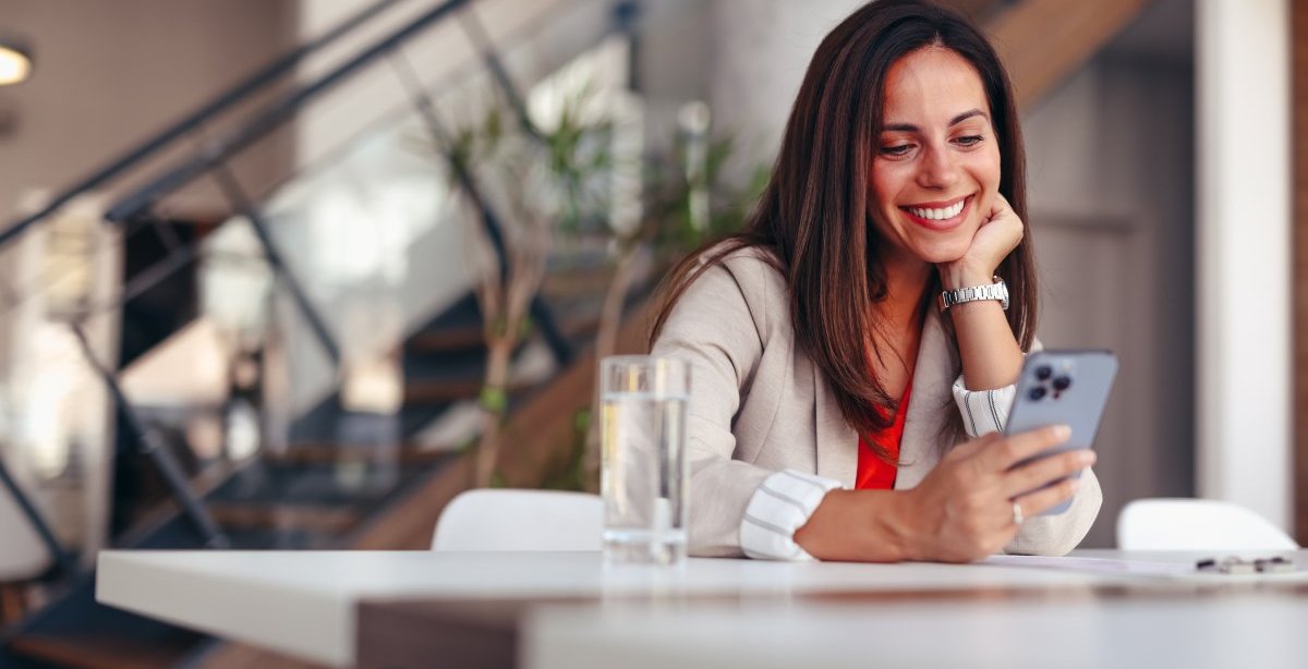 A imagem mostra uma mulher em uma casa usando um celular. Ela está apoiada em uma mesa com um copo de água à frente e olha para o smartphone após ter sido impactada por alguma publicação que fez uso de impulsionamento com a ajuda do TikTok Ads.