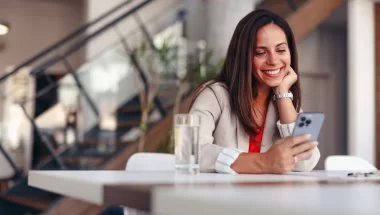 A imagem mostra uma mulher em uma casa usando um celular. Ela está apoiada em uma mesa com um copo de água à frente e olha para o smartphone após ter sido impactada por alguma publicação que fez uso de impulsionamento com a ajuda do TikTok Ads.