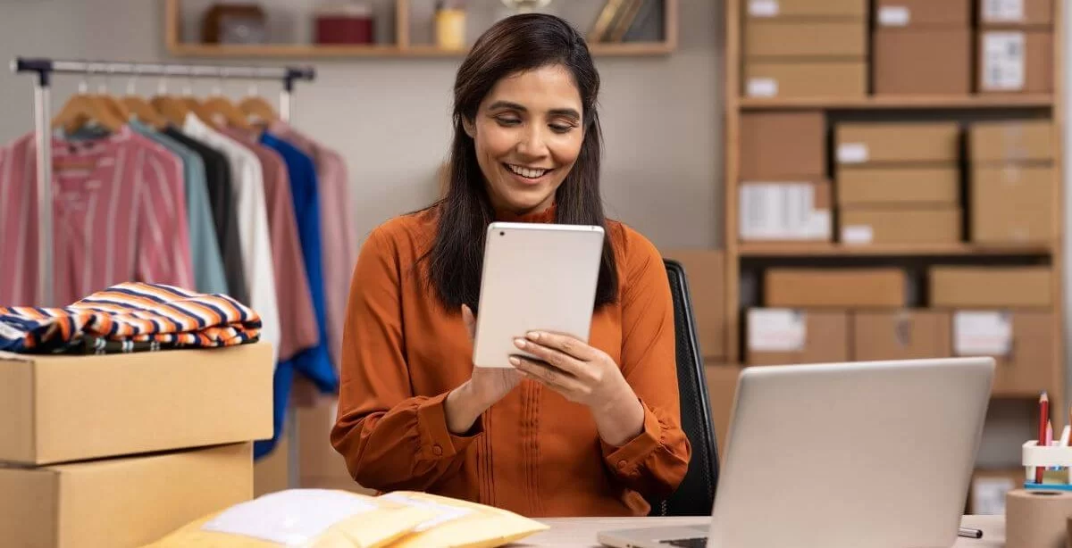 A imagem mostra uma mulher no estoque de uma loja de roupas mexendo em um tablet. Ela também está diante de um notebook com dois pacotes, possivelmente procurando produtos para revenda online.