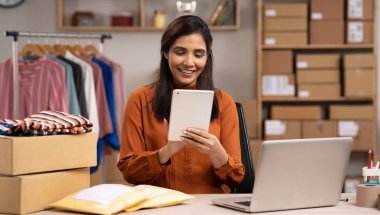 A imagem mostra uma mulher no estoque de uma loja de roupas mexendo em um tablet. Ela também está diante de um notebook com dois pacotes, possivelmente procurando produtos para revenda online.