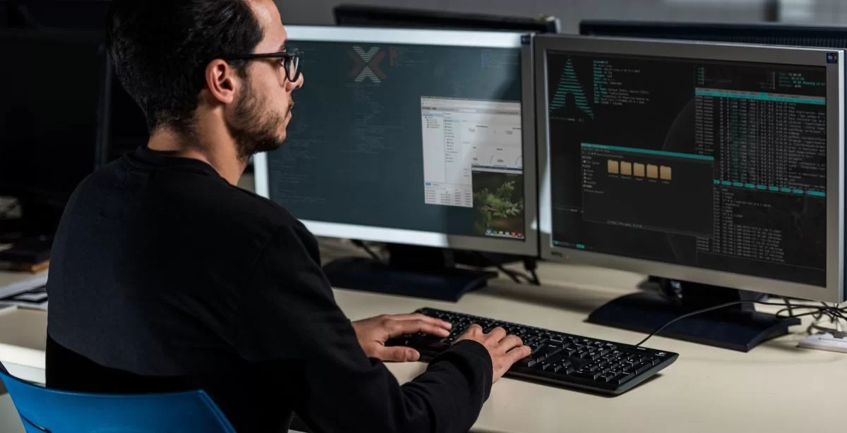 A imagem mostra um homem jovem em frente à mesa de trabalho dele. Ele está digitando no teclado enquanto verifica linhas de código geradas com o auxílio do SCP Linux.