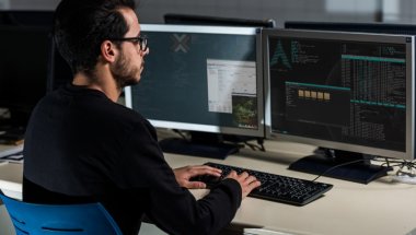 A imagem mostra um homem jovem em frente à mesa de trabalho dele. Ele está digitando no teclado enquanto verifica linhas de código geradas com o auxílio do SCP Linux.