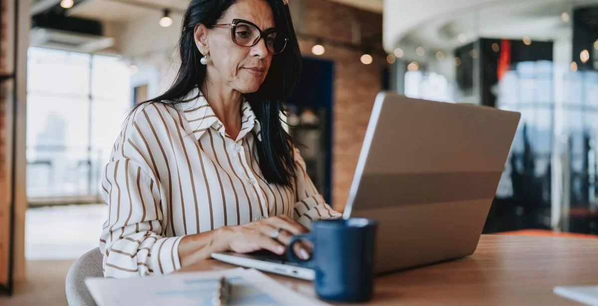 A imagem mostra uma mulher trabalhando em um escritório. Ela está digitando em um notebook o endereço de um site confiável, checando com cuidado todos os elementos para não cair em nenhum tipo de golpe.